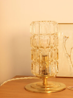 Table lamp placed on a wooden bedside table. The lamp is composed of a vintage moulded and chiselled glass globe resting on a brushed brass base.