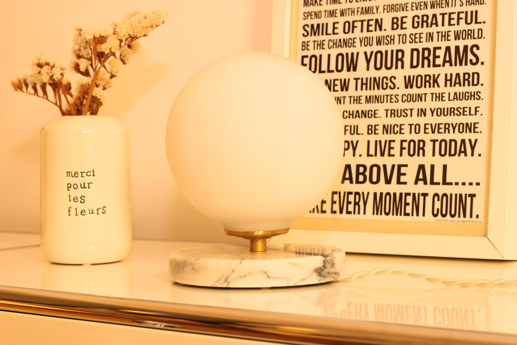 The lamp rests on a white metal piece of furniture. A frame is placed behind the opaline lamp with its white marble base and grey veins. A cream-colored flower pot is also part of the scene, featuring a text in French. Dried flowers are in the flower pot.