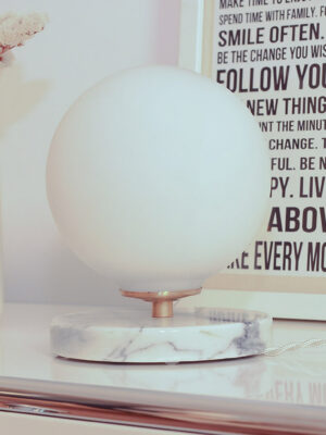 The lamp rests on a white metal piece of furniture. A frame is placed behind the opaline lamp with its white marble base and grey veins. A cream-colored flower pot is also part of the scene, featuring a text in French. Dried flowers are in the flower pot.