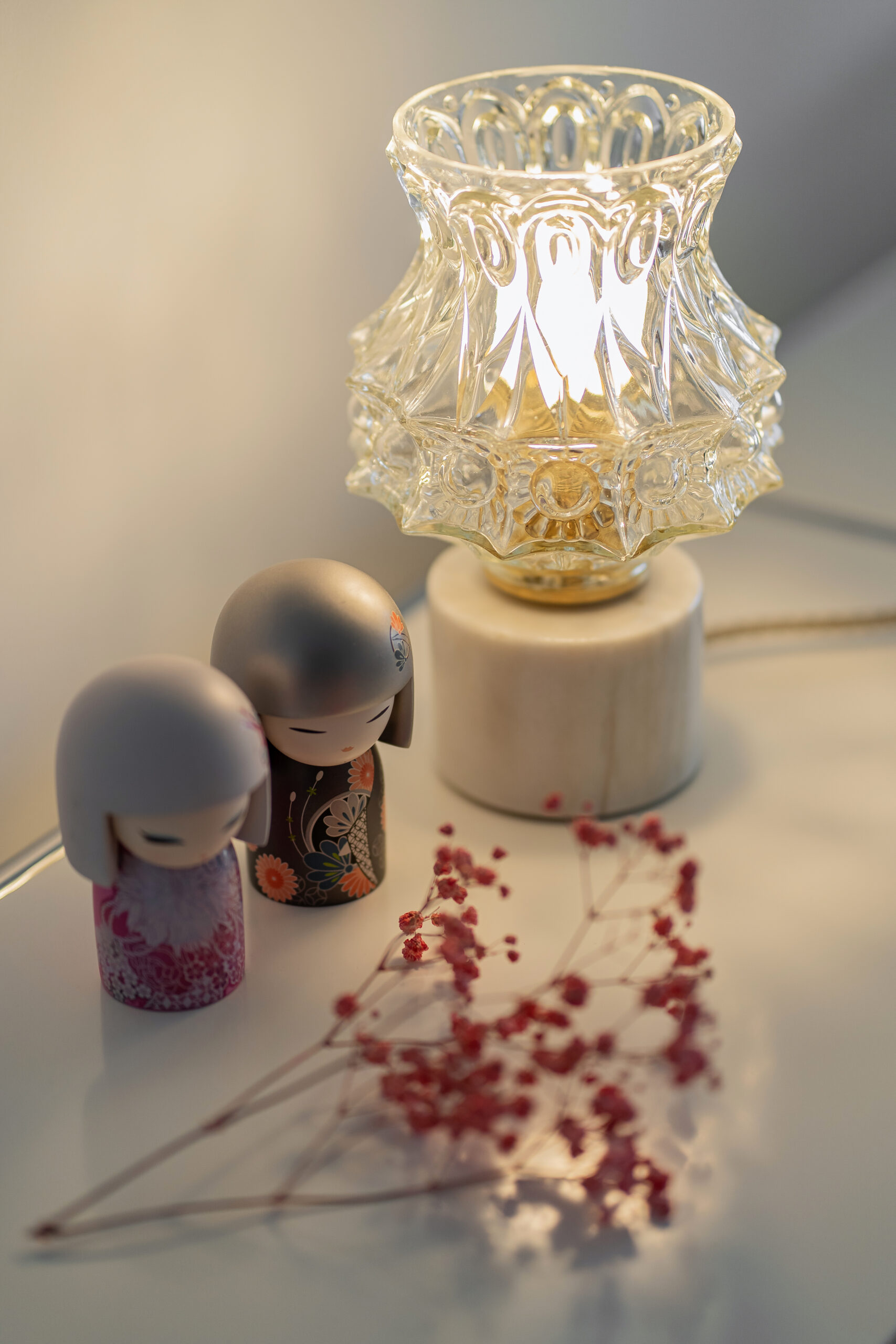 Candeeiro vintage de vidro com base em mármore, iluminado sobre uma mesa branca, ao lado de duas pequenas figuras japonesas e flores secas.