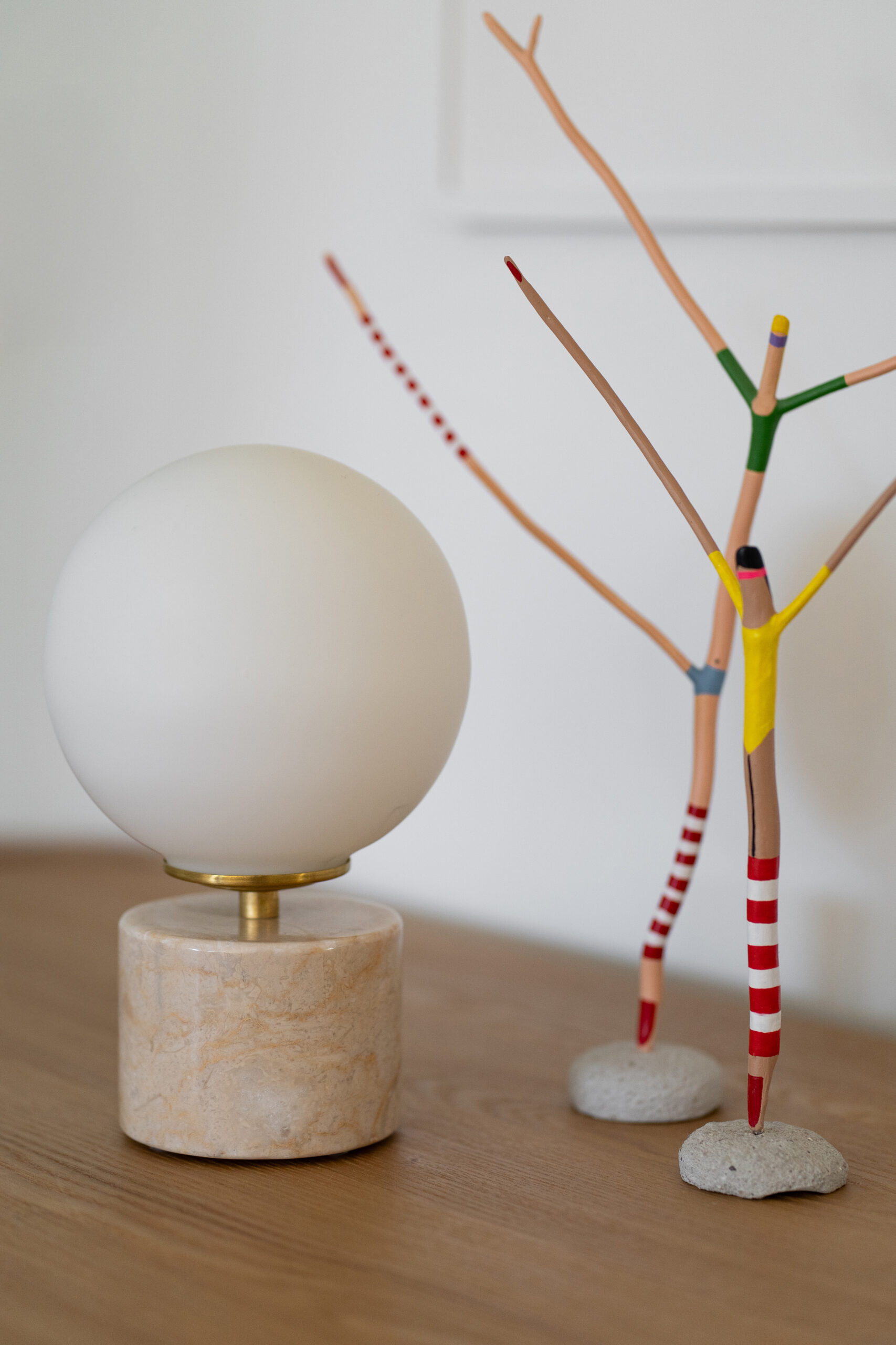 Candeeiro de design com cúpula redonda em vidro opalino e base em mármore bege, sobre um móvel de madeira junto a uma escultura decorativa colorida.