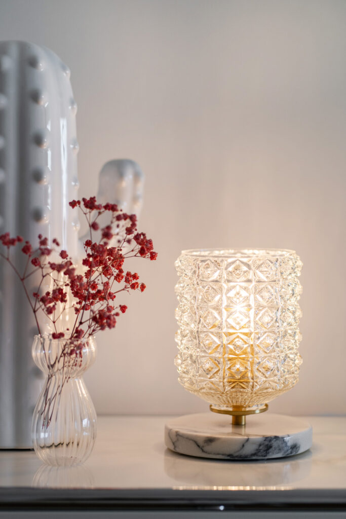 Candeeiro decorativo de vidro lapidado com base em mármore e latão, iluminado sobre um móvel branco, ao lado de um vaso de vidro com pequenas flores secas vermelhas.