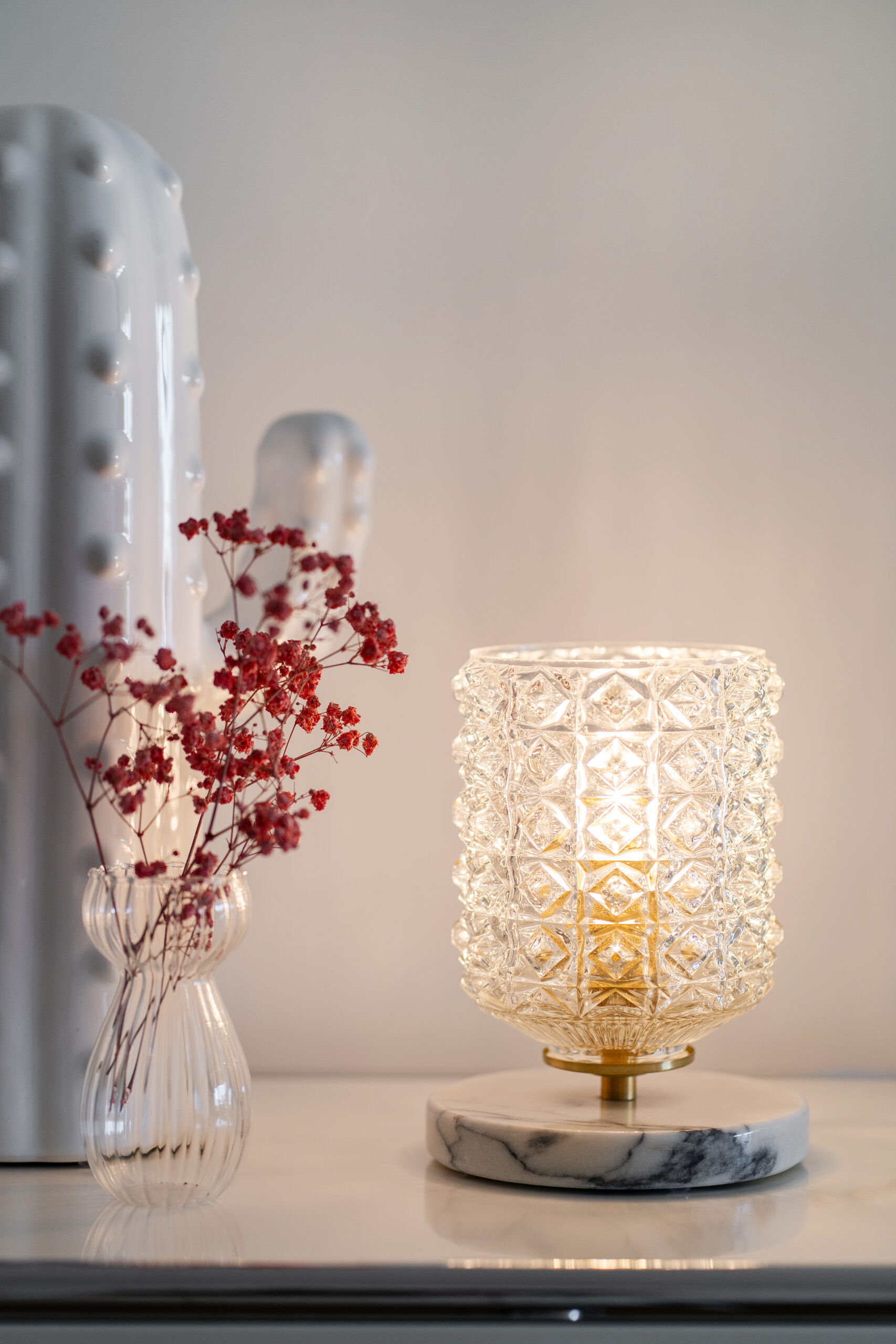 Candeeiro decorativo de vidro lapidado com base em mármore e latão, iluminado sobre um móvel branco, ao lado de um vaso de vidro com pequenas flores secas vermelhas.