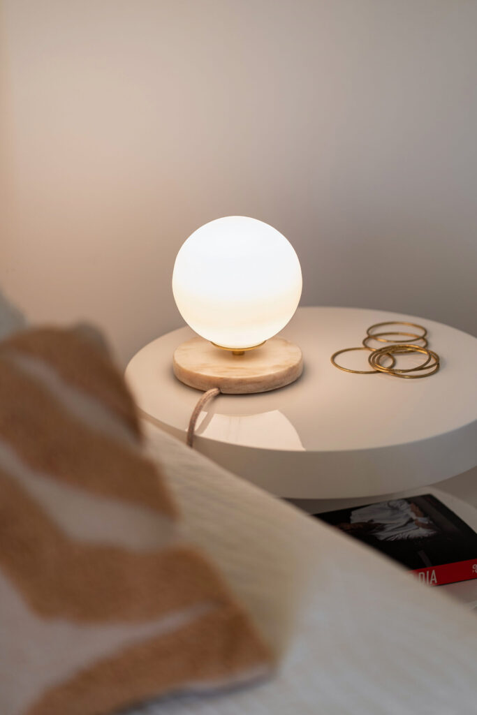 Isabelle opaline glass lamp lit on a round marble bedside table with beige veining, with a few gold bracelets next to it.