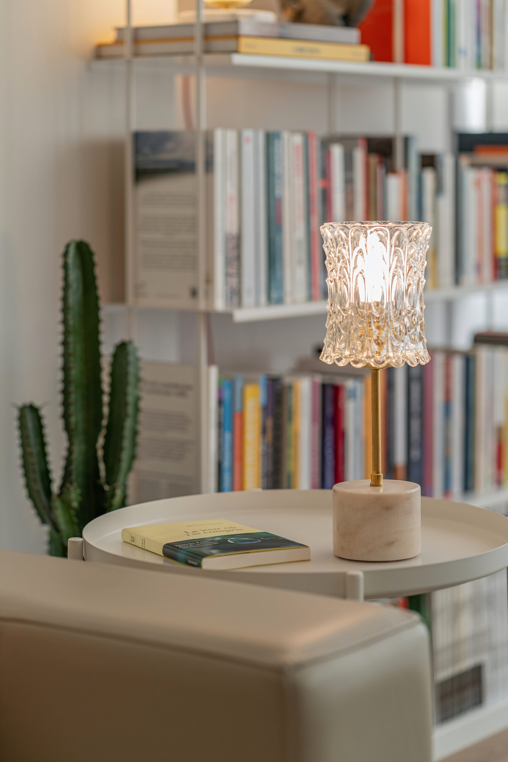 Candeeiro decorativo de vidro lapidado com base em mármore e latão, aceso sobre uma mesa redonda branca em frente a uma estante, com um livro ao lado e um cato ao fundo.