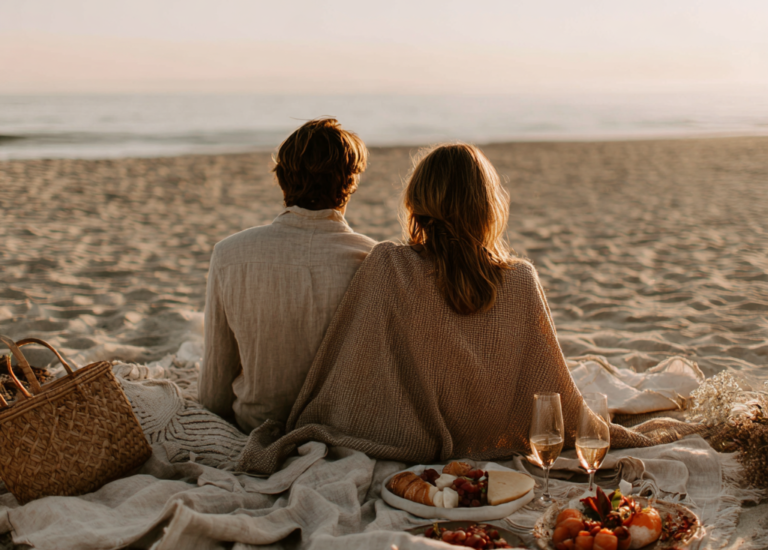 Couple sitting on the beach at sunset, sharing a romantic picnic under a cozy blanket, warm and intimate atmosphere.