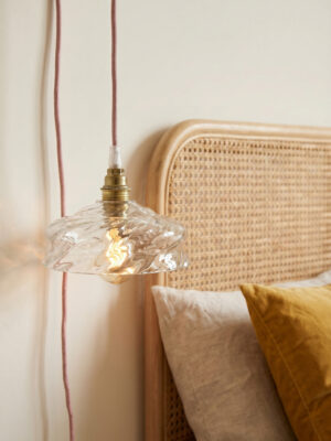 Vista aproximada da suspensão Cloé em vidro moldado transparente ondulado e latão envelhecido acesa com lâmpada de filamento, cordão de algodão rosa visível, cabeceira em palhinha natural e almofada de veludo ocre em segundo plano