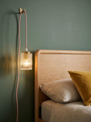 Suspensão Julia em vidro moldado-prensado cilíndrico transparente com motivo ponta de diamante e caneluras verticais, latão polido, acesa e pendurada num gancho de latão numa parede verde salva profundo, reflexos dourados na parede, ao lado de uma cama com cabeceira em carvalho claro, almofada de veludo ocre e almofada de linho bege, cordão de algodão rosa