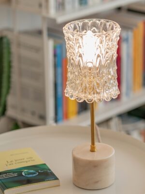 Candeeiro de mesa Carlota com base cilíndrica em mármore bege, haste em latão e globo em vidro moldado vintage com motivo floral sobre uma mesa diante de uma estante