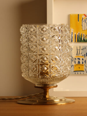 Candeeiro de mesa vintage Céleste da Alfama Chic aceso — globo em vidro moldado-prensado com motivos diamante sobre base em latão dourado polido, colocado num móvel de madeira