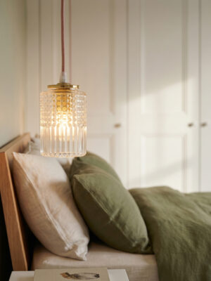 Grande plano da suspensão Julia em vidro moldado-prensado cilíndrico transparente com motivo ponta de diamante em cima e caneluras verticais em baixo, latão polido, acesa, cordão de algodão rosa, cama em madeira com almofadas em linho verde oliva e bege e armário branco com painéis em segundo plano desfocado