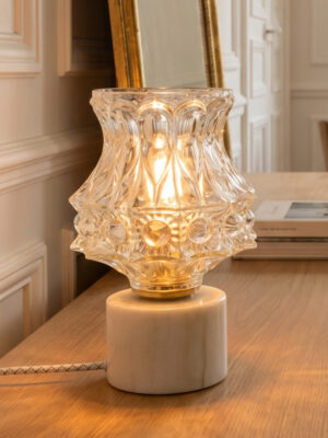 Lampe décorative vintage Angèle d'Alfama Chic allumée en plan serré sur un meuble en bois naturel dans un salon haussmannien, globe en verre moulé-pressé forme évasée en corolle à motifs organiques diffusant une lumière dorée et chaude, bague en laiton doré, base cylindrique en marbre d'Estremoz veiné beige et vert (Alentejo), câble torsadé blanc, cadre de miroir doré et boiseries en arrière-plan, assemblée à la main à Lisbonne