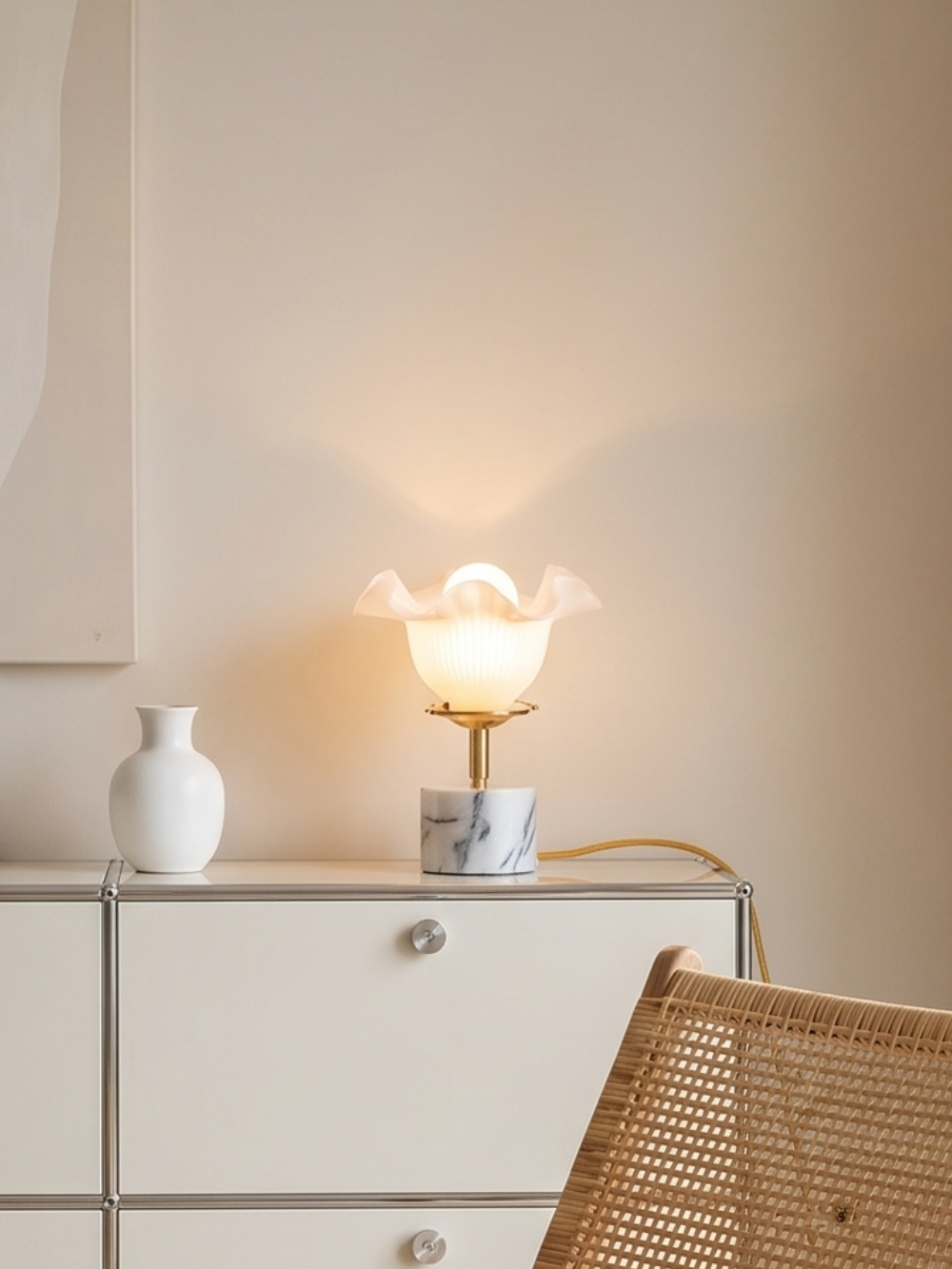 Lampe tulipe vintage Hortense d'Alfama Chic allumée sur un meuble bas blanc, globe opaline dépoli à bords ondulés projetant un halo de lumière chaude sur le mur beige, base cylindrique en marbre d'Estremoz veiné gris et blanc, monture laiton doré, câble torsadé ocre, vase blanc en céramique à côté, chaise en rotin au premier plan, assemblée à la main à Lisbonne