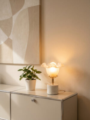 Lampe tulipe vintage Hortense d'Alfama Chic allumée sur un meuble bas blanc, globe opaline à bords ondulés diffusant une lumière dorée et chaude, base cylindrique en marbre d'Estremoz, monture laiton doré, plante verte et tableau abstrait en arrière-plan, assemblée à la main à Lisbonne