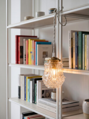 Vintage hand lamp Louise switched on hanging in a white bookshelf, mould-pressed glass globe with floral pattern, white twisted cord, coloured books in background — Alfama Chic