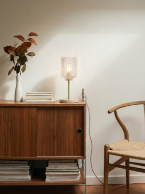 Vintage desk lamp Rose by Alfama Chic lit on a dark wood sideboard in a warm vintage-style living room, cylindrical cut glass globe with geometric patterns diffusing warm golden light, Lioz stone base (Sintra region), slender gilded brass stem, red twisted textile cord, white vase with rust-coloured leafy branch and stacked books beside it, wooden chair in the foreground, hand-assembled in Lisbon