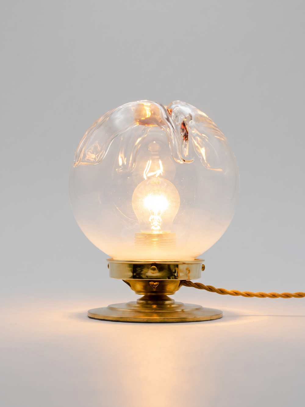 Lampe à poser vintage globe verre style Murano transparent allumée, base laiton massif et câble torsadé doré, fond gris neutre