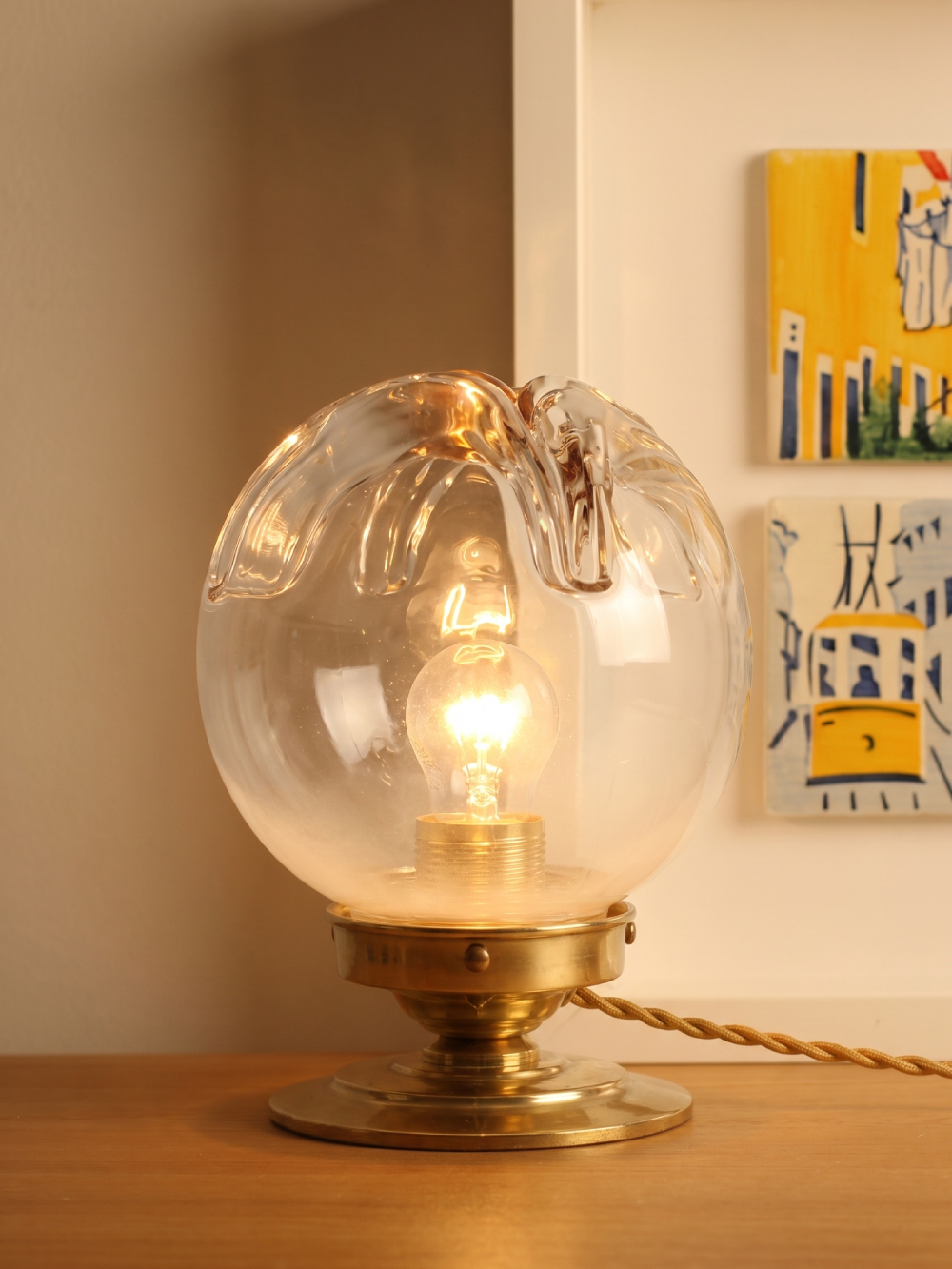 Lampe vintage globe verre style Murano allumée sur meuble en bois, base laiton massif, câble torsadé doré, tableaux colorés en arrière-plan