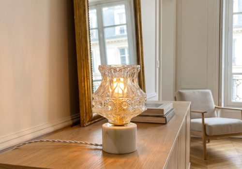 Candeeiro decorativo vintage Angèle da Alfama Chic aceso sobre um móvel buffet em madeira natural numa sala haussmanniana elegante em tons bege e creme, globo em vidro moldado a pressão forma evasada em corolha a difundir uma luz quente e dourada, base cilíndrica em mármore de Estremoz com veios bege e verde (Alentejo), cabo têxtil torcido branco, grande espelho com moldura dourada e poltrona em linho bege ao fundo, soalho em espinha de peixe, montado à mão em Lisboa